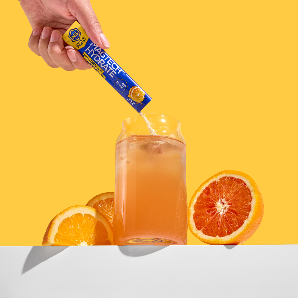 Hand pouring a drink mix into a glass with oranges on a yellow background
