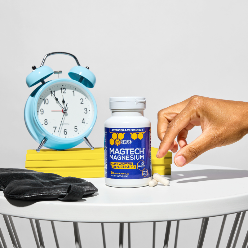 Hand holding a supplement next to a bottle of Magtech Magnesium, an alarm clock, and a container on a white surface.