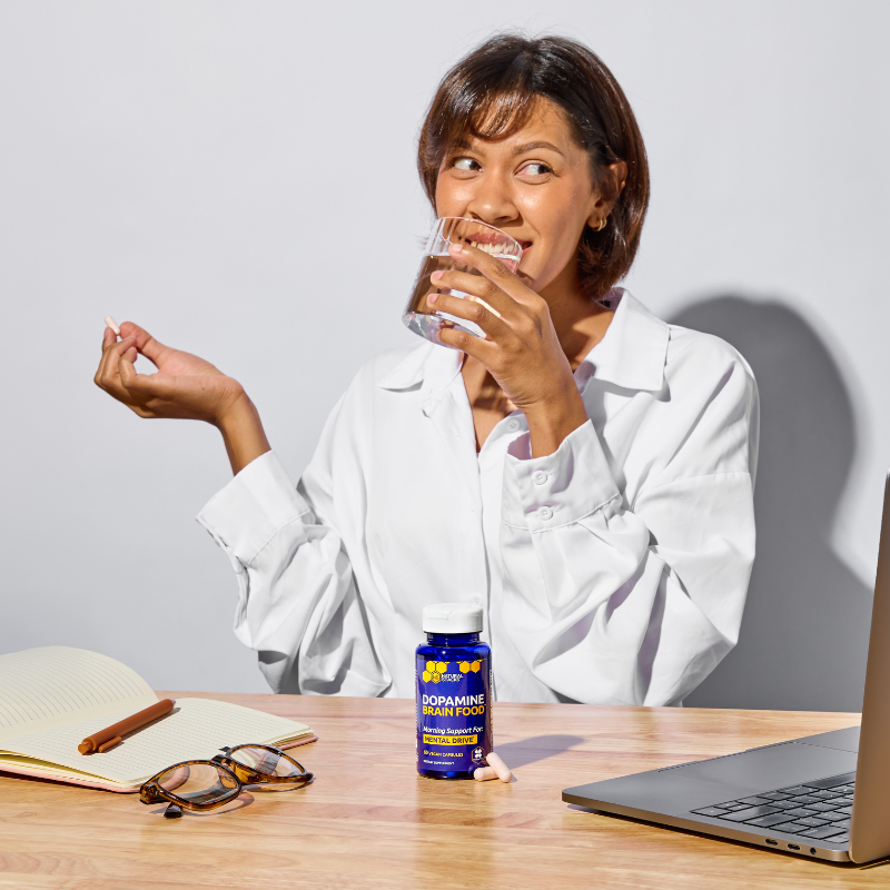 Woman in a white shirt drinking water at a desk with a bottle of Gummy Bearz Dopaamine Boosters.
